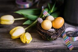 Osternest mit Eiern, buntem Geschenkband und gelben Tulpen auf rustikaler Holzunterlage