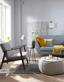 A pouffe in front of a sofa with cushions, a side table and a retro floor lamp against a wallpapered piece of chipboard