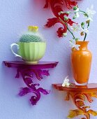 Bunte kleine Wandkonsolen mit orangefarbener Vase, Orchideenzweig und Kaktus in nostalgischer Tasse dekoriert