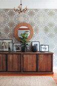 Framed photos on solid-wood sideboard below round mirror on wall with op-art wallpaper