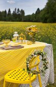 Wreath of flowers on yellow metal chair next to table set in yellow colour scheme with field of flowering rapeseed in background