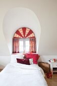 Bed below arched dormer window with semi-circular transom and decorative curtains in shades of red
