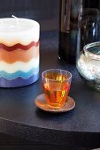 Orange gefärbtes Schnapsglas auf Holzuntersetzer neben einer Kerze