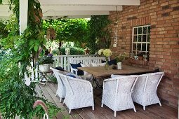 White wicker armchairs and broad table on pleasant wooden terrace between brick façade and climber-covered pergola supports