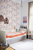 Grey and white wooden bed with storage drawers and orange fitted sheet against patterned wallpaper in child's bedroom