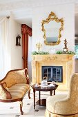 Comfortable, elegant seating area with antique seating and coffee table in front of fireplace below gilt-framed mirror