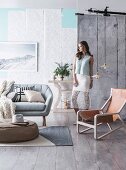Round upholstered table on woven carpet, sofa and Scandinavian armchair with leather seat, woman against wall with wallpaper strips