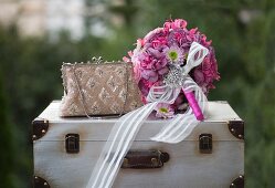 Romantic, pink bridal bouquet with white ribbon and brooch next to elegant handbag on vintage suitcase