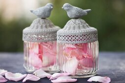 Two vintage-style glass jars with bird figurines on lids filled with pink rose petals