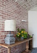 Rustikaler Konsolentisch mit Tischleuchte und Blumentrauss vor Backsteinwand