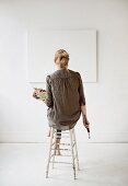 Künstlerin in Atelier mit Pinseln und Malerpalette auf dem Hocker vor einer weissen Leinwand