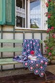 Crocheted blanket of hexagons in shades of blue and purple on vintage garden bench against rustic façade of house