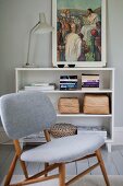 Chair with pale grey upholstery in front of retro table lamp and religious painting on half-height shelving unit
