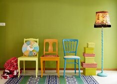Colourful wooden chairs, patchwork cushion, stack of boxes and vintage standard lamp against line green wall