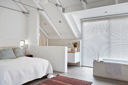 Schlafzimmer im weißen Dachgeschoss mit offenem Bad, freistehende Badewanne vor Fenstern und geschlossener Jalousie