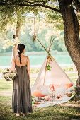 Woman wearing long summer dress holding basket of flowers in front of romantic picnic in teepee
