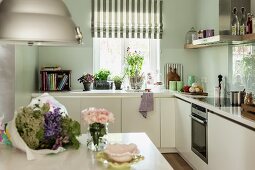 weiße Einbauküche mit Blumenstrauss auf Tisch, Übereck-Küchenzeile vor Fenster mit Küchenutensilien