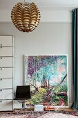 Modern artwork leant against pale grey wall behind vintage swivel chair and metallic pendant lamp