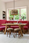Button-tufted red bench below window and rustic dining table below owl-patterned pendant lamp in corner of dining room