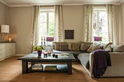 Couchtisch und Eckpolstersofa vor Fenstern in elegantem, beigefarbenem Wohnraum