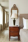 Bedside lamp on antique, marquetry bedside cabinet and mirror in bedroom with corridor along one side
