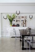 Colonial-style table, postmodern Ghost chairs, pictures and antlers above antique chest of drawers
