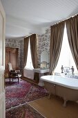 Cosy, vintage-style ensuite bathroom with free-standing bathtub, draped beige curtains, various Oriental rugs and toile de jouy wallpaper