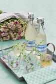 Gläser und Limonadenflaschen mit Schnappverschluss im Vintage Stil auf schlichtem Metall Tablett, im Hintergrund üppiger Rosenstrauss