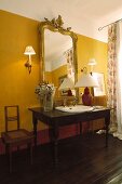 Washstand made from desk with integrated sink and elegant, gilt-framed mirror on yellow-painted wall