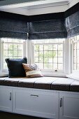Cushioned window seat with cupboards below, lead glazed bay window and dark grey Roman blinds
