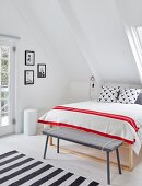 Simple bench at foot of double bed with red and white striped blanket below sloping ceiling