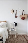 Stacked books on white wooden chair with hat on backrest below bags and hat hung on wall-mounted pegs in bedroom