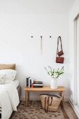 Stack of books and vase of flowering branches on wooden bedside table below bags hanging from coat pegs