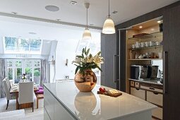 Glossy, white island counter and fitted cupboards with open sliding doors in elegant, open-plan kitchen