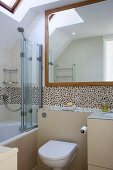 Bathroom with toilet against pale beige half-height wall below mosaic tiles; bathtub with glass screen to one side