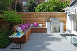 Purple cushions on fitted wooden bench and barbecue on sunny terrace with modern screen