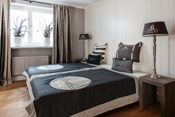 Doppelbett mit bedruckter anthrazitfarbener Tagesdecke und modernen Nachttischen, Tischleuchten vor weisser, holzverkleideter Wand