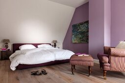 Double bed with white bed linen, comfortable armchair with matching footstool and purple-painted accent wall in attic room