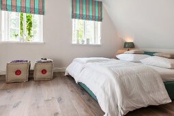 Bedroom in modern rustic style; double bed with white bed linen under sloping ceiling and pouffes below striped Roman blinds on windows