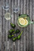 Slices of lemon in glass vase of lemonade, drinking glasses and fresh green figs on wooden surface