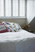 Pale bedspread and embroidered cushions on bed below window with closed interior shutters