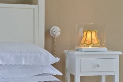 Lit, vintage bedside lamp in glass case on white bedside table with drawer
