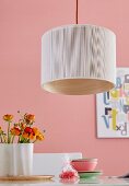 A pendent lamp with light fabric over bent wooden veneer over a table laid with buttercups against a pink wall
