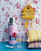 Pink thermos flask with floral motif next to brightly striped cushions below yellow cuckoo clock on rose-patterned wallpaper