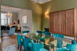 Oval glass table, petrol-blue upholstered chairs and modern cupboard in grand dining room with view into living room through wide, open doorway