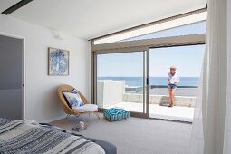 Airy bedroom with window front and sea view, woman on glass balustrade on terrace