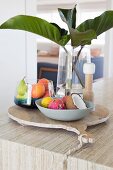 Still life with tropical fruits in fruit bowls next to glass vase with green leaves