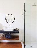Verglaste Duschkabine neben rundem Wandspiegel und Waschbecken auf Waschtischplatte
