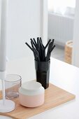 Black drinking straws, china pot, glass and candlestick on wooden chopping board