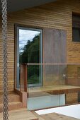 Fassadenausschnitt mit Holzverkleidung, Glastür und Metallplatte, davor Glasbrüstung und Treppenstufen